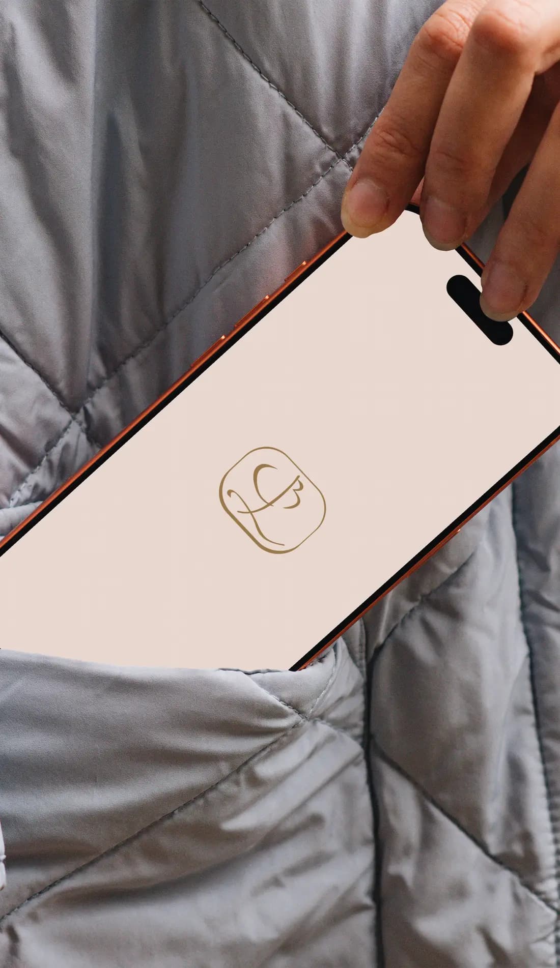 A hand slipping a phone into the pocket of a quilted grey jacket; the screen shows the gold BL monogram on rose sand