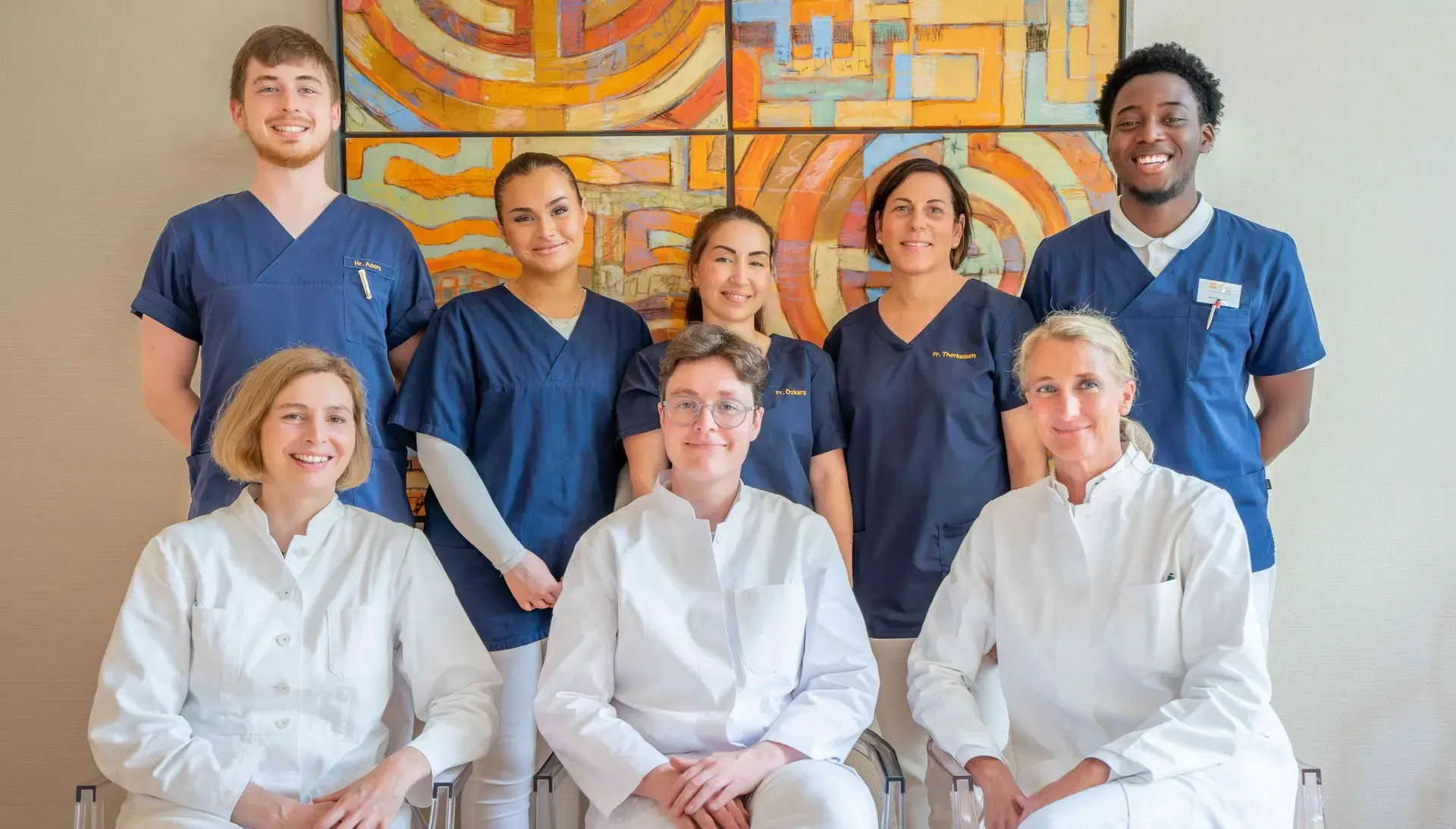 The HNO in Eppendorf team, three ENT doctors in white coats with their practice staff in navy scrubs, in front of a colourful painting in the practice