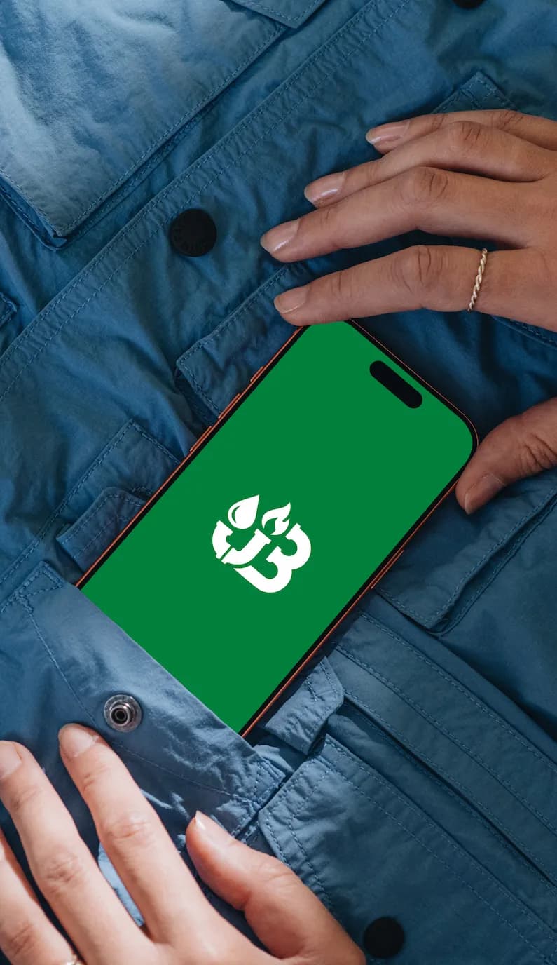 Hands slipping a phone into the pocket of a blue cotton jacket; the screen shows the white JB drop-and-flame mark on green